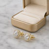 Gold earrings with clear gemstones on a marble surface next to an open beige jewelry box.